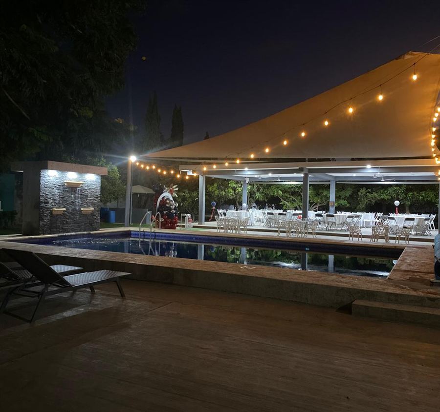 El salón de noche, con las mesas montadas bajo la velaria iluminada y la alberca al frente.